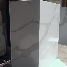 Dawid Szablewski - Biały, kwadratowy blok z szarymi żyłkami, prawdopodobnie z kamienia lub konglomeratu, stoi na metalowym stole w warsztacie kamieniarskim. Widoczne okno w tle.