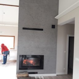 Dawid Szablewski - Nowoczesny kominek z szarą ścianą o fakturze betonu, płomienie w palenisku, drewno ułożone obok. W tle widoczna osoba w kuchni, co dodaje wnętrzu życia.