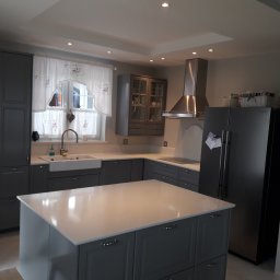 Dawid Szablewski - Szara kuchnia z wyspą i białym blatem, okapem ze stali nierdzewnej i oknem z firanką w kwiaty. Nowoczesne oświetlenie sufitowe.