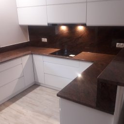 Dawid Szablewski - Nowoczesna kuchnia z blatem z ciemnego kamienia, białymi szafkami i brązowym panelem nad blatem. Widoczne gniazdka elektryczne i płyta indukcyjna.