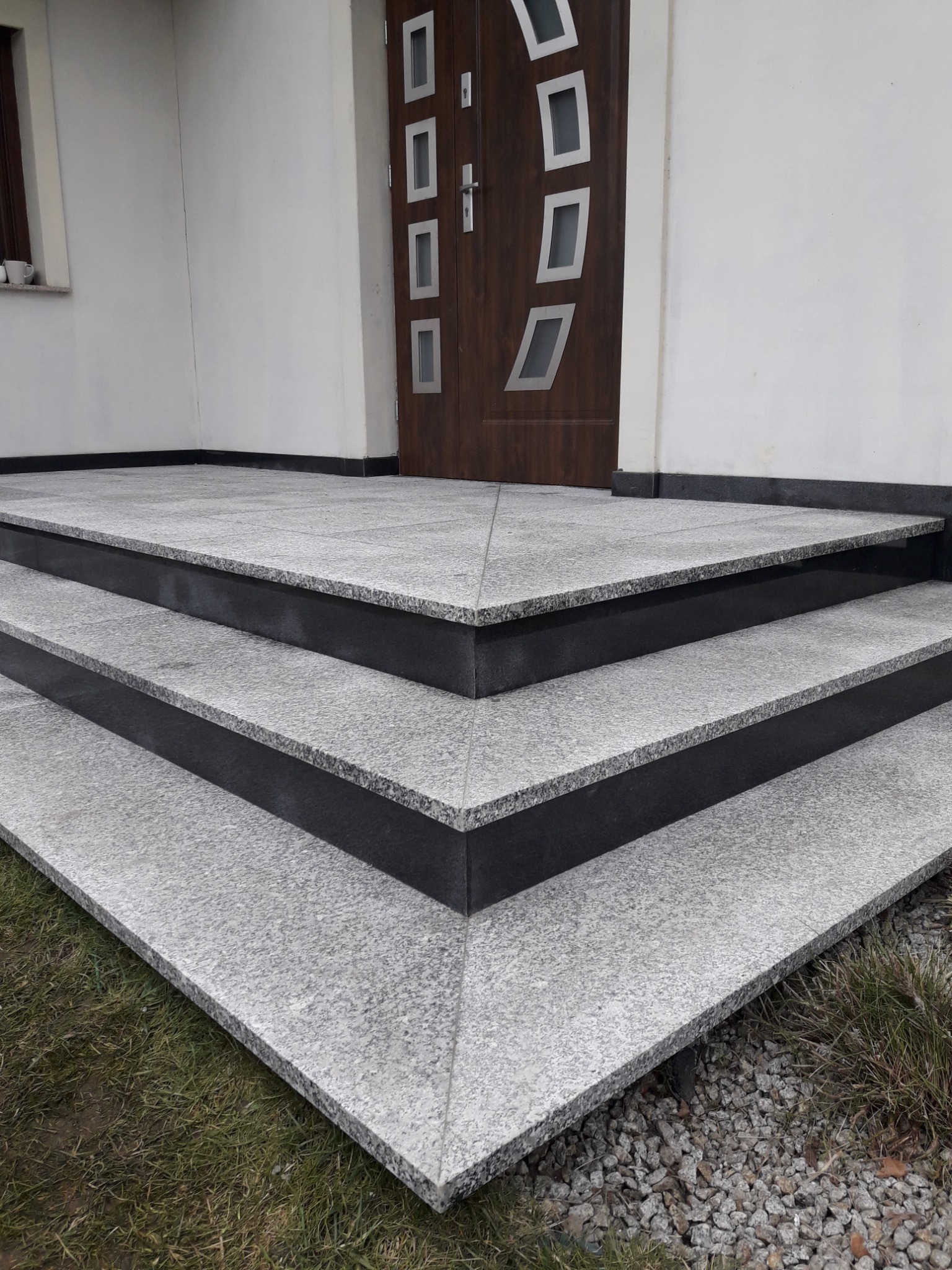 Granitowe schody wejściowe z czarnymi podstopnicami prowadzą do domu. Widoczna część drzwi wejściowych z oryginalnymi przeszkleniami.