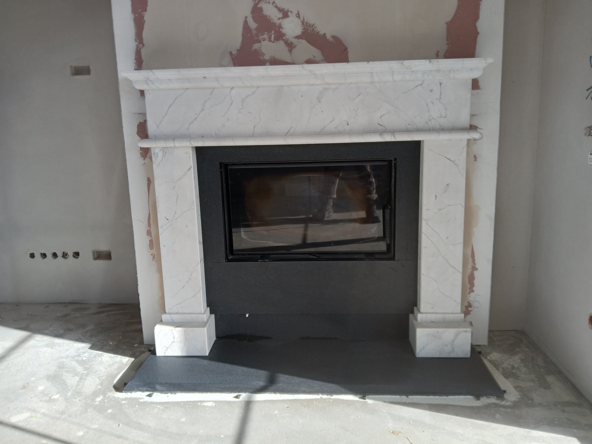 Marmurowy portal kominkowy z czarnym wkładem, w surowym wnętrzu. Widoczne ślady po pracach wykończeniowych na ścianach. Minimalistyczny design.