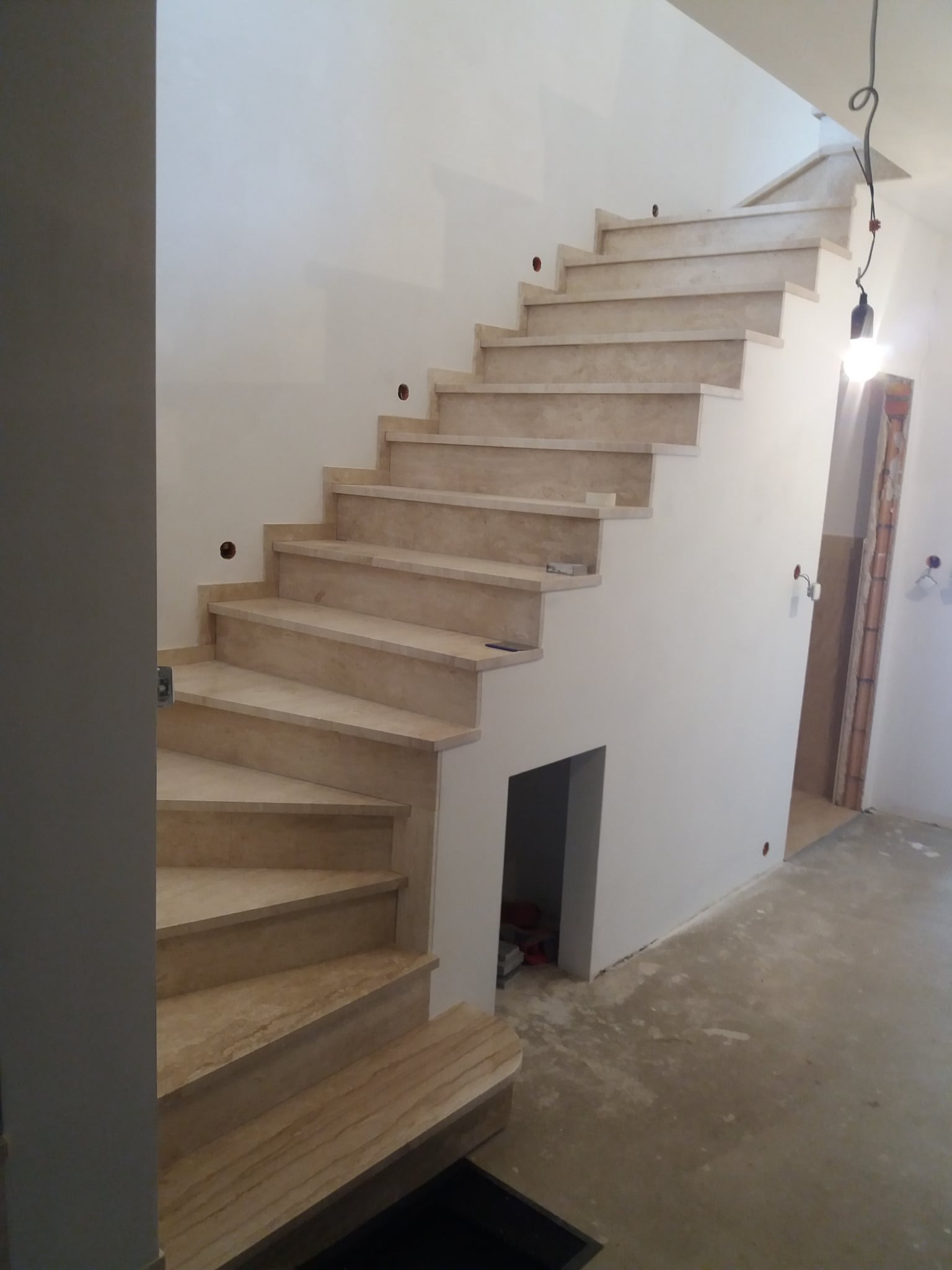 Jasne, marmurowe schody wewnętrzne z podstopnicami, w trakcie montażu. Widoczne otwory na gniazdka elektryczne w ścianie. Surowy beton na podłodze.