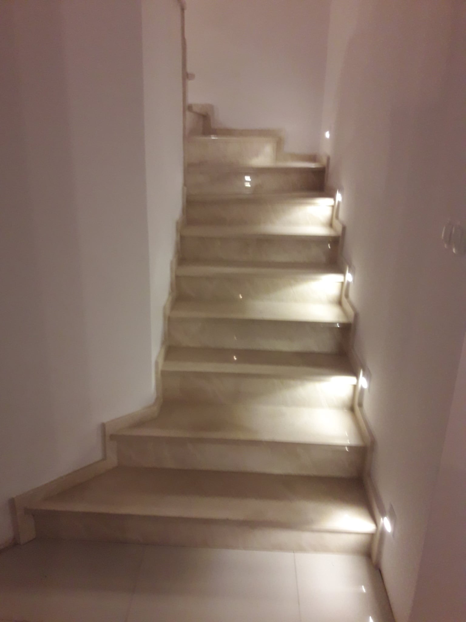Kamienne schody wewnętrzne z podświetleniem LED wzdłuż ściany, widok z dołu. Beżowe stopnie i podstopnice tworzą elegancki wygląd.