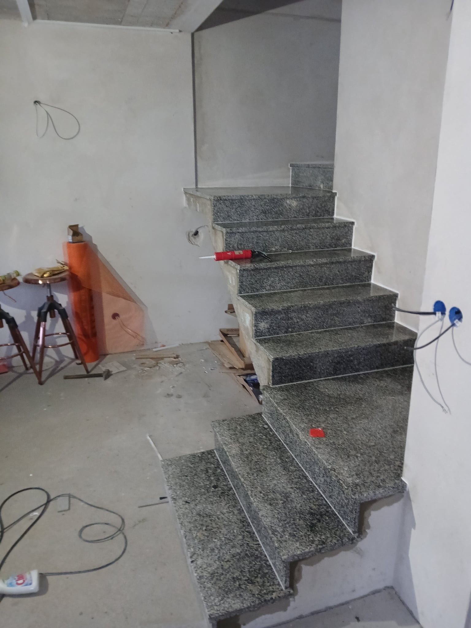 Granitowe schody wewnętrzne w trakcie montażu, widoczne narzędzia i materiały budowlane. Surowe ściany w tle. Detal: czerwony aplikator do silikonu na schodach.
