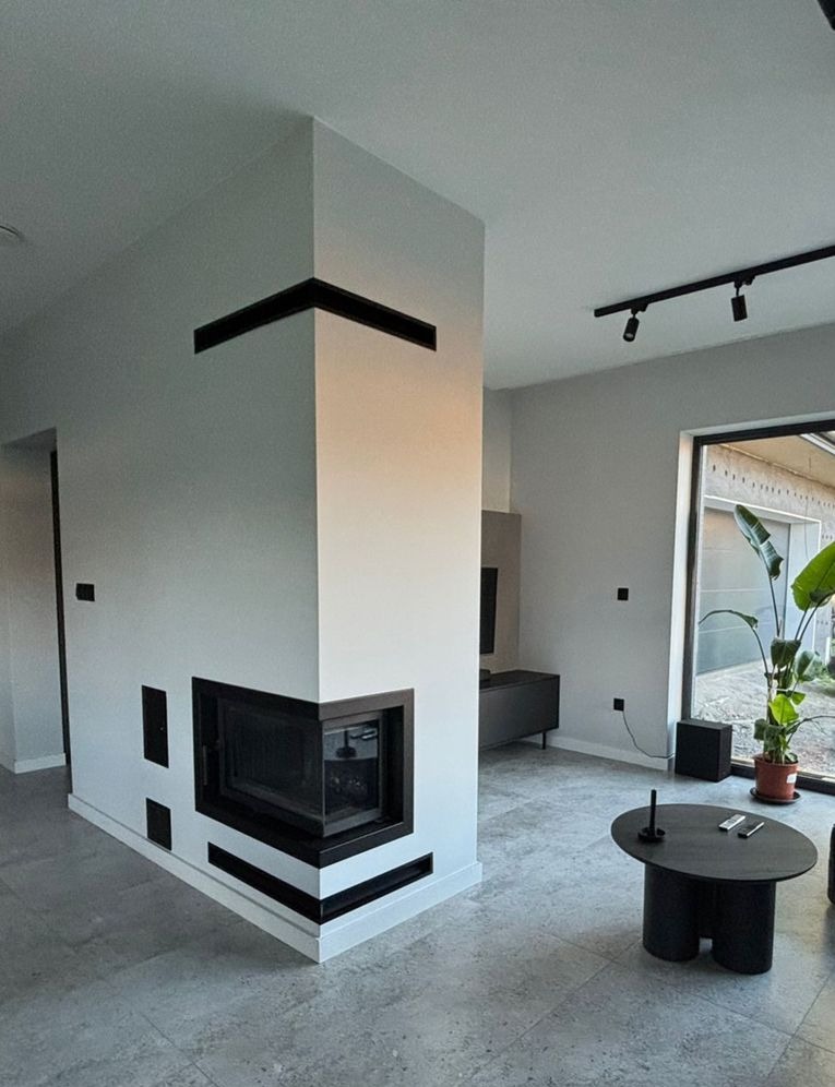 Nowoczesny kominek narożny z czarnymi akcentami w minimalistycznym salonie. Widoczne detale wykończenia i oświetlenie punktowe.