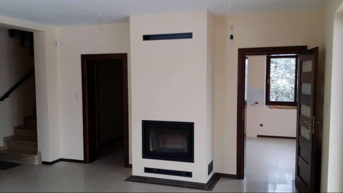 Nowoczesny kominek z czarnym wkładem, w jasnym salonie z widocznymi schodami i otwartymi drzwiami. Minimalistyczny design, neutralna kolorystyka ścian i podłóg.