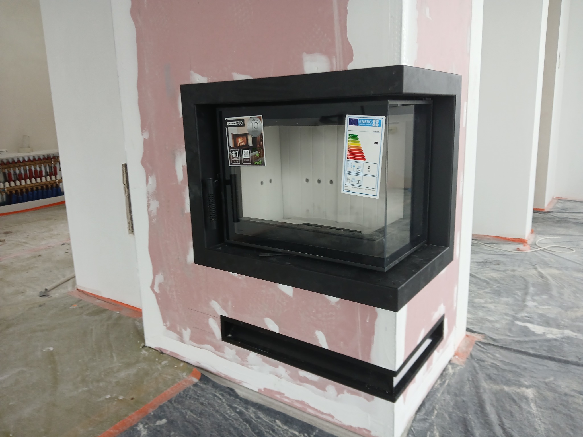 Nowoczesny kominek z czarną obudową w trakcie montażu, widoczne naklejki informacyjne na szybie, surowy stan wykończenia ścian i podłogi, instalacja CO w tle.