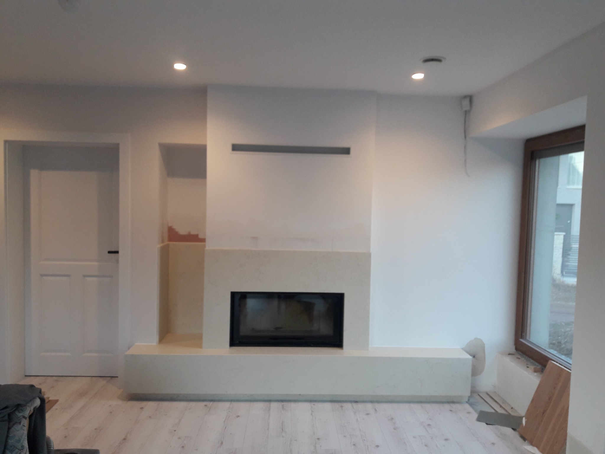 Nowoczesny kominek z jasnego kamienia w minimalistycznym salonie. Prosta forma, subtelne oświetlenie punktowe, drewniana podłoga. Widok na okno i drzwi.