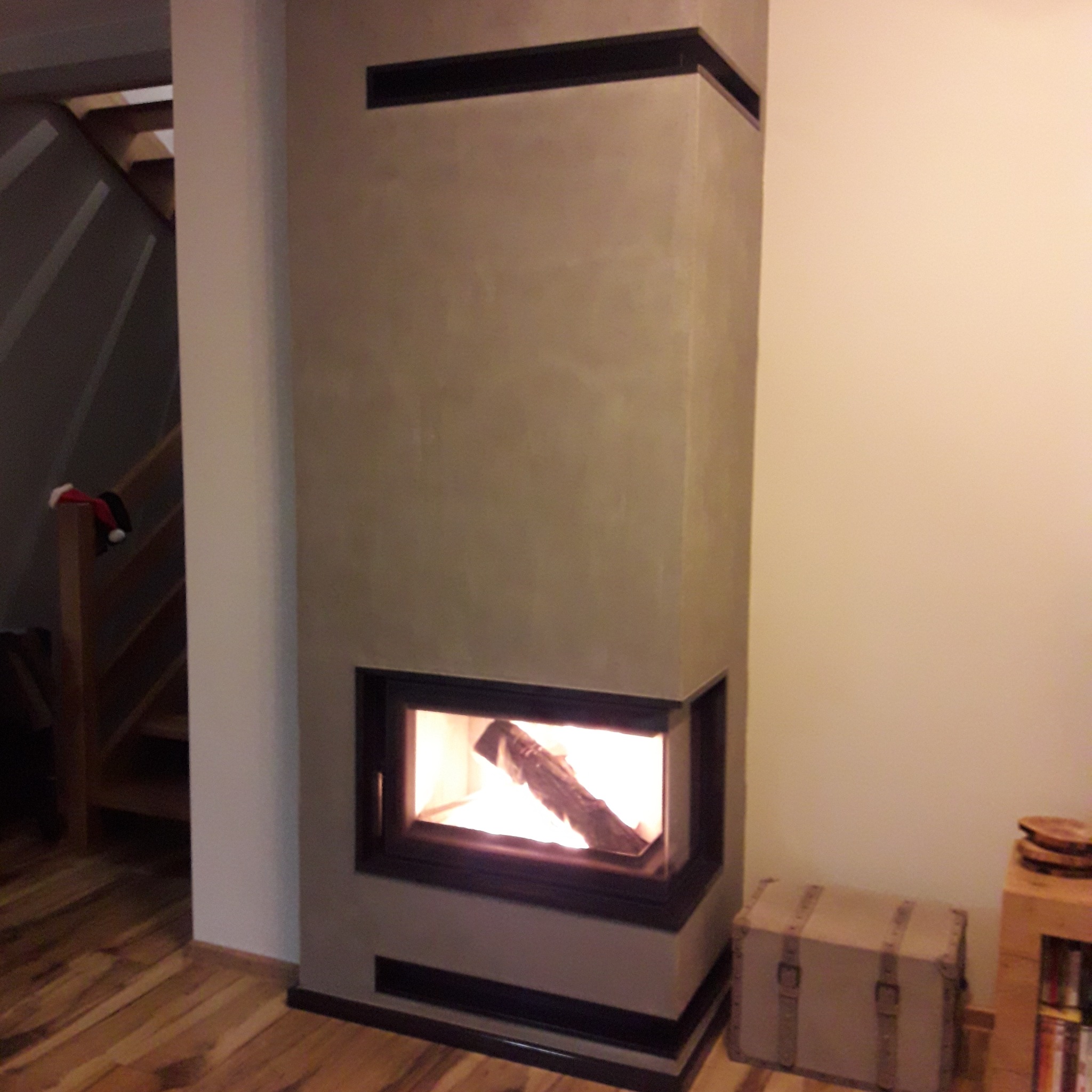 Nowoczesny kominek narożny z paleniskiem, obudowany beżowym tynkiem strukturalnym z czarnymi akcentami. W tle drewniane schody i skrzynia.