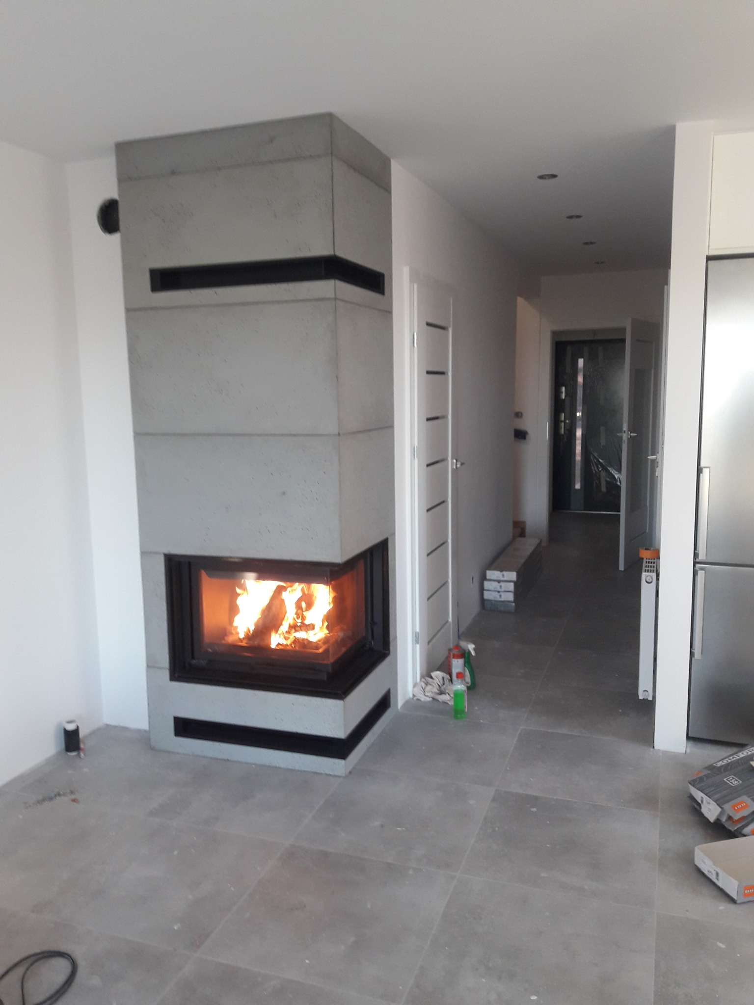 Nowoczesny kominek z szarego betonu architektonicznego, z widocznym płomieniem, w minimalistycznym wnętrzu z szarą podłogą i białymi ścianami. Widok na korytarz.