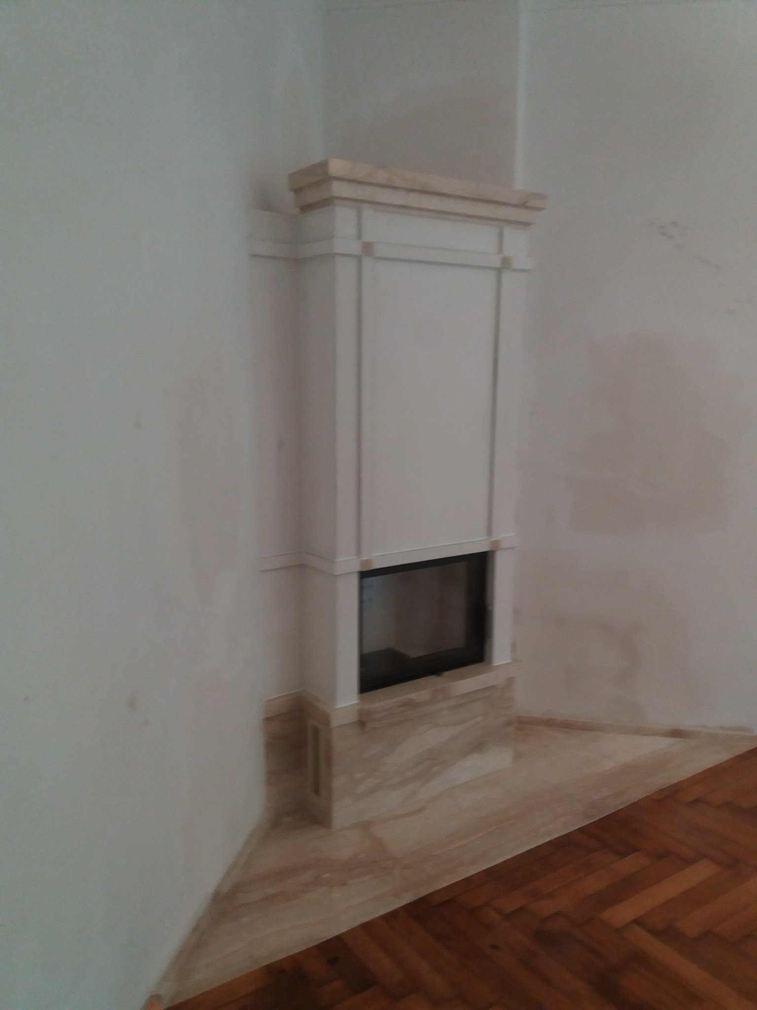 Narożny kominek z białym obramowaniem i marmurowym wykończeniem podstawy, wkomponowany w jasne wnętrze z drewnianą podłogą. Widok z boku, oświetlenie dzienne.