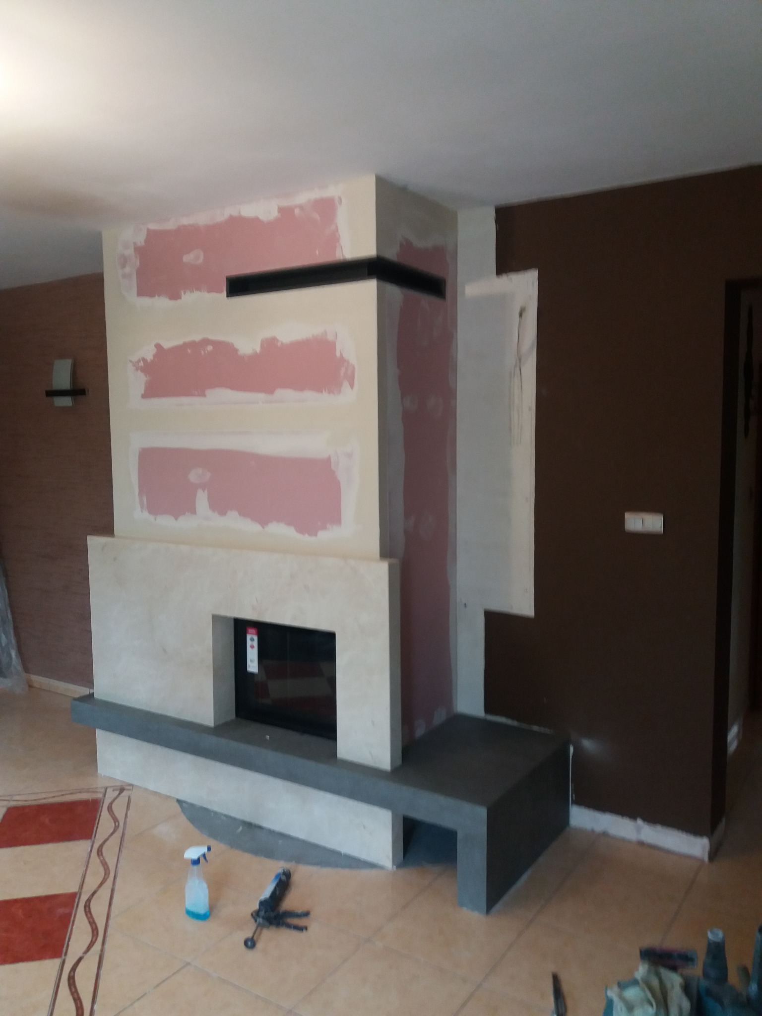 Wnętrze z kominkiem w trakcie wykończenia. Widoczne płyty gipsowe na ścianie, obudowa kominka z jasnego kamienia i ciemny cokół. Na podłodze narzędzia i butelka z płynem.