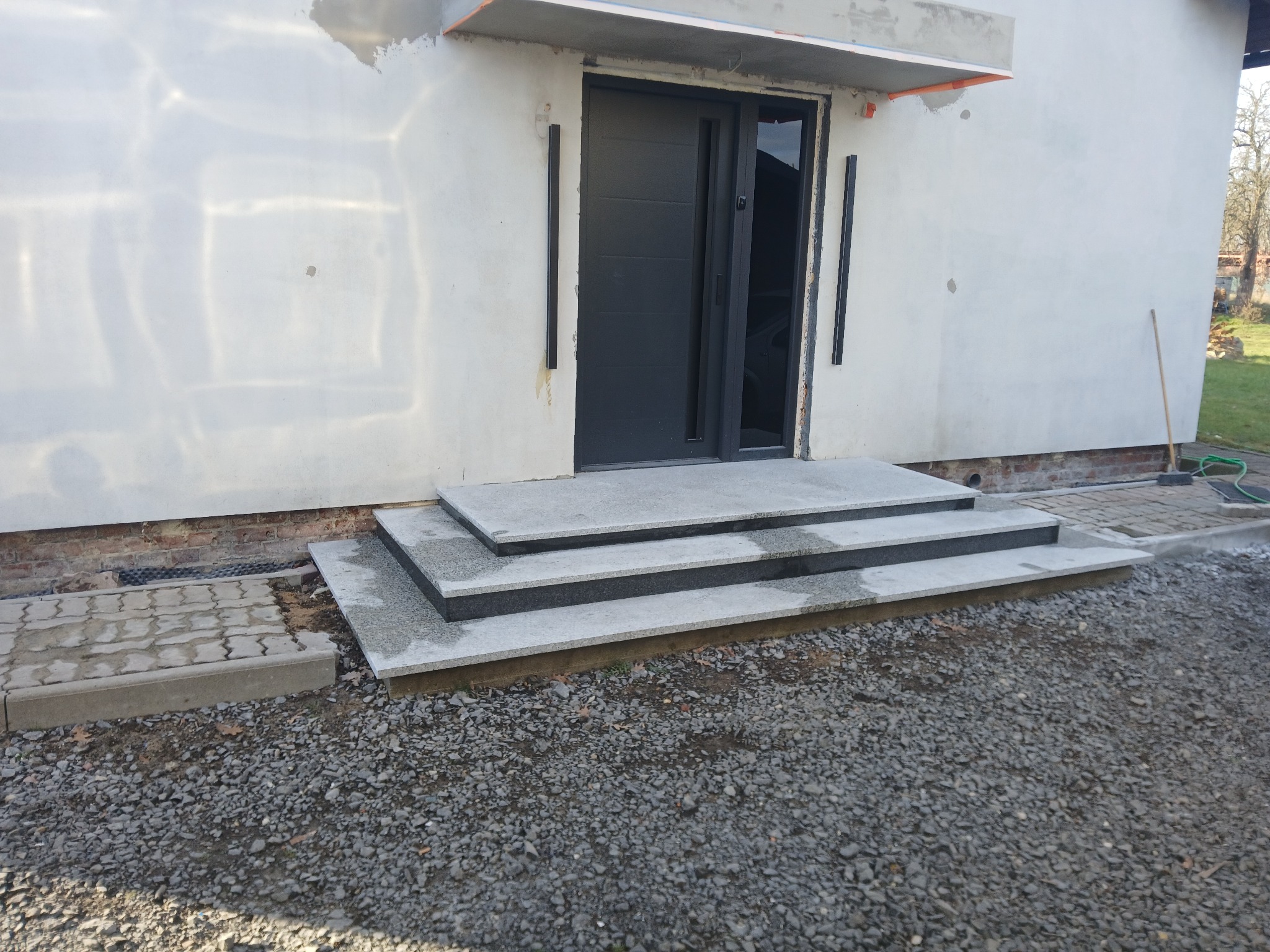 Granitowe schody wejściowe z trzema stopniami prowadzące do nowoczesnych, czarnych drzwi wejściowych w jasnym budynku. Widoczne szare kamienie na podłożu.