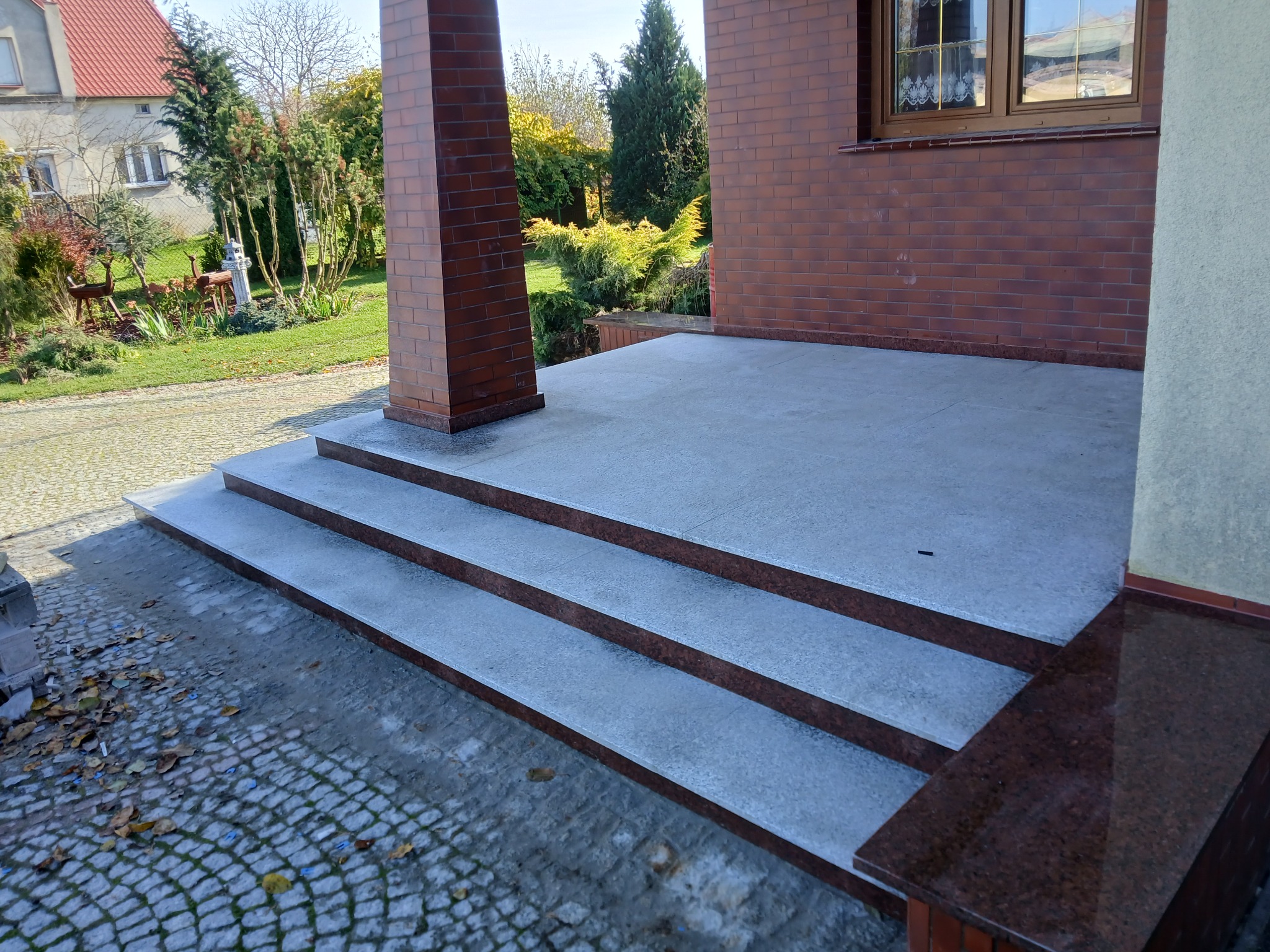 Zewnętrzne schody wejściowe z jasnoszarego kamienia z ciemnobrązowymi krawędziami, prowadzące do domu z cegły. Widoczna kostka brukowa i fragment ogrodu.
