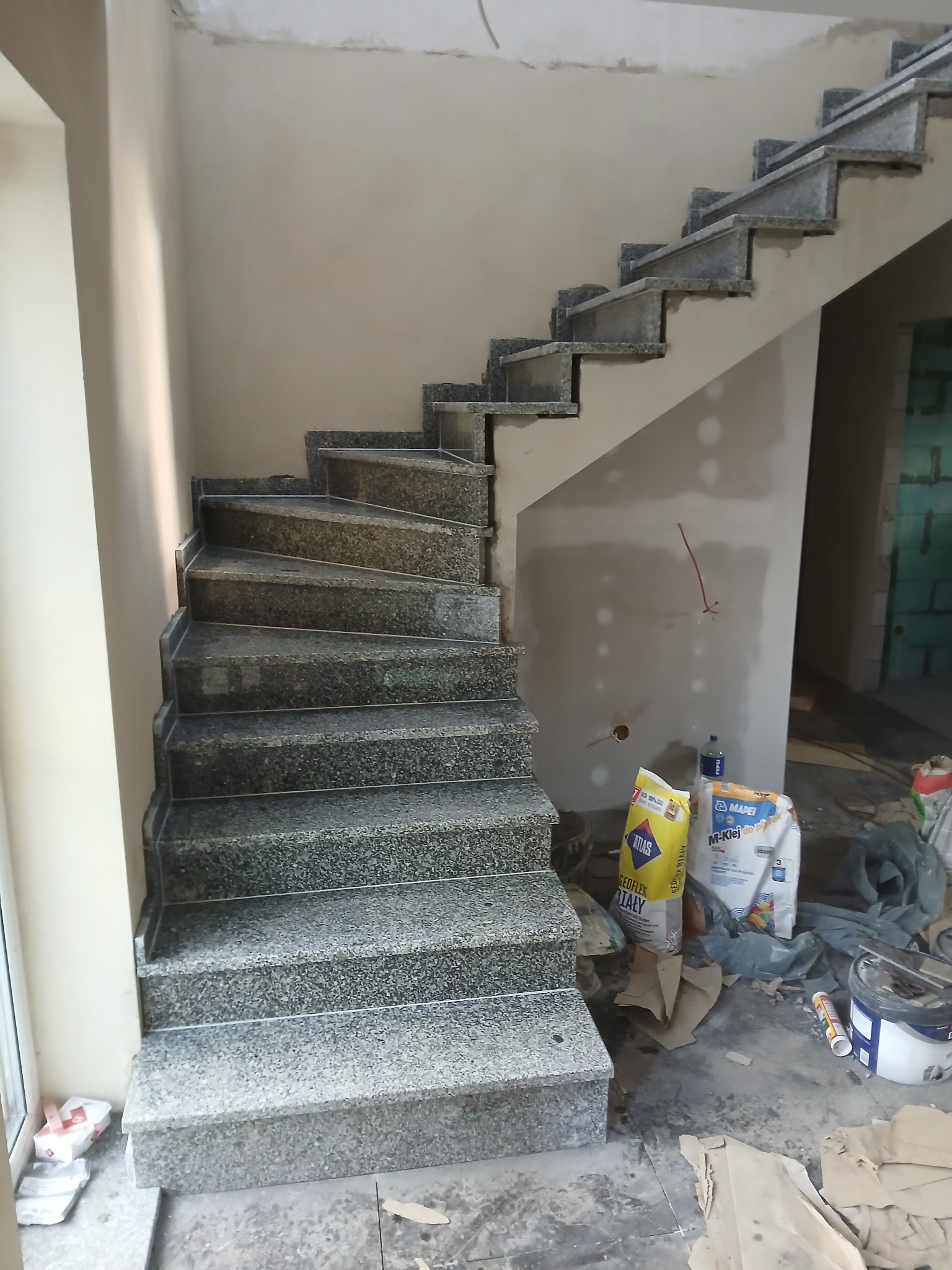 Granitowe schody spiralne w trakcie montażu, widoczne worki z klejem i narzędzia. Surowe ściany w tle. Ujęcie z dołu, podkreślające kształt i fakturę kamienia.