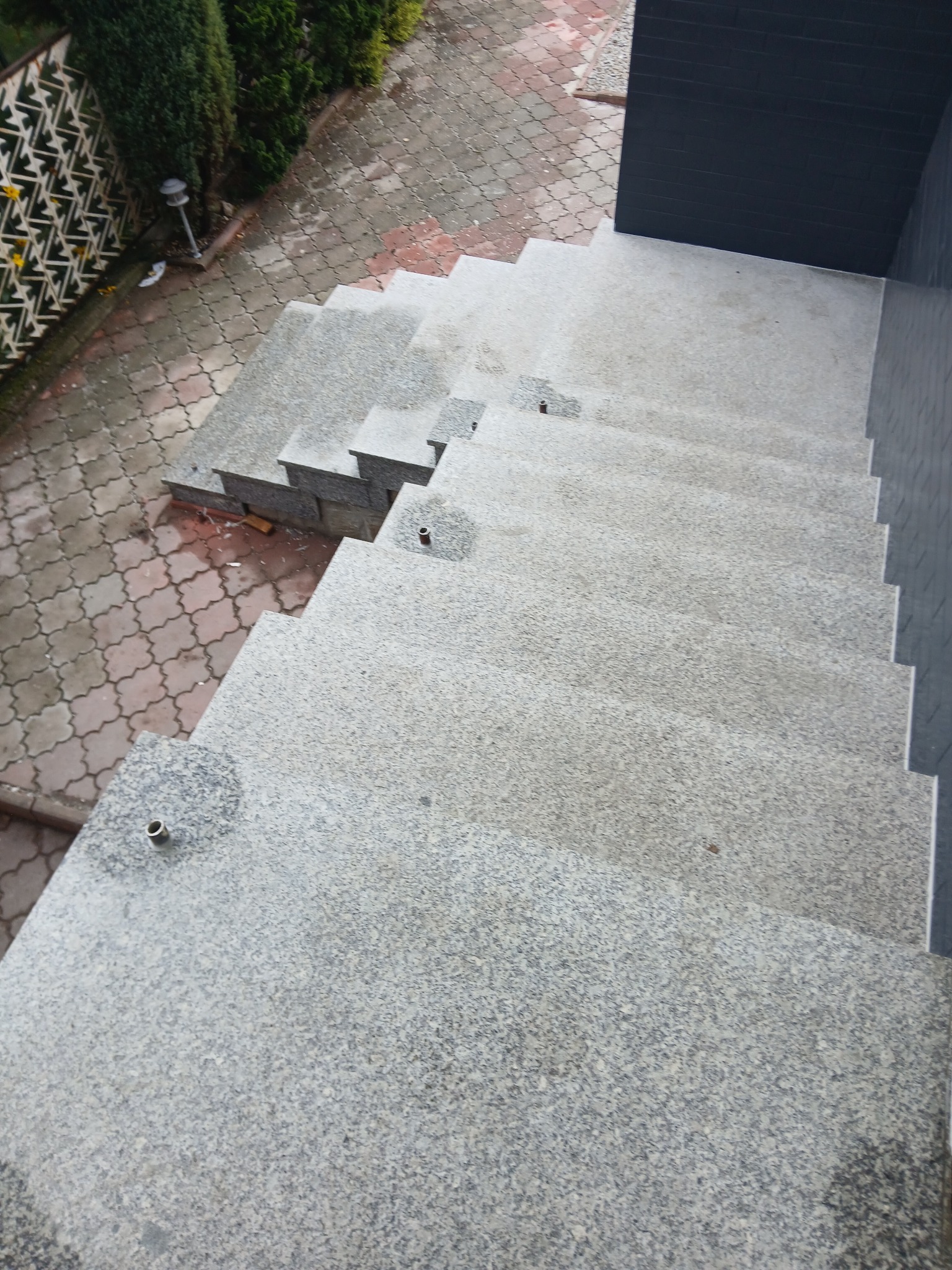 Zewnętrzne schody z jasnoszarego kamienia z metalowymi elementami, widok z góry, obok kostka brukowa i ciemna ściana budynku. Minimalistyczny design.