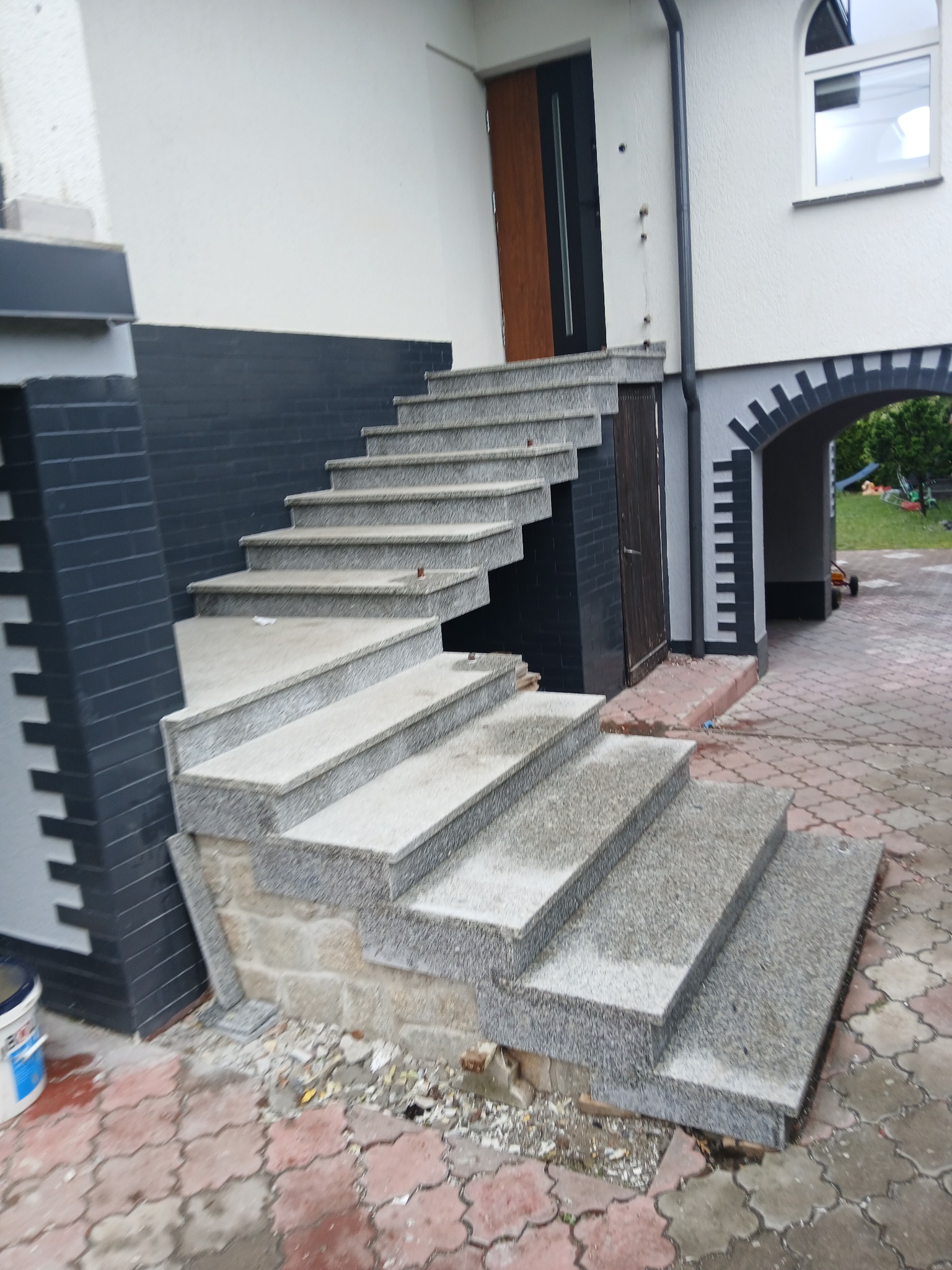 Zewnętrzne schody z szarego kamienia prowadzące do drzwi wejściowych, z widoczną kostką brukową i elementami wykończeniowymi budynku w tle. Ujęcie z dołu.