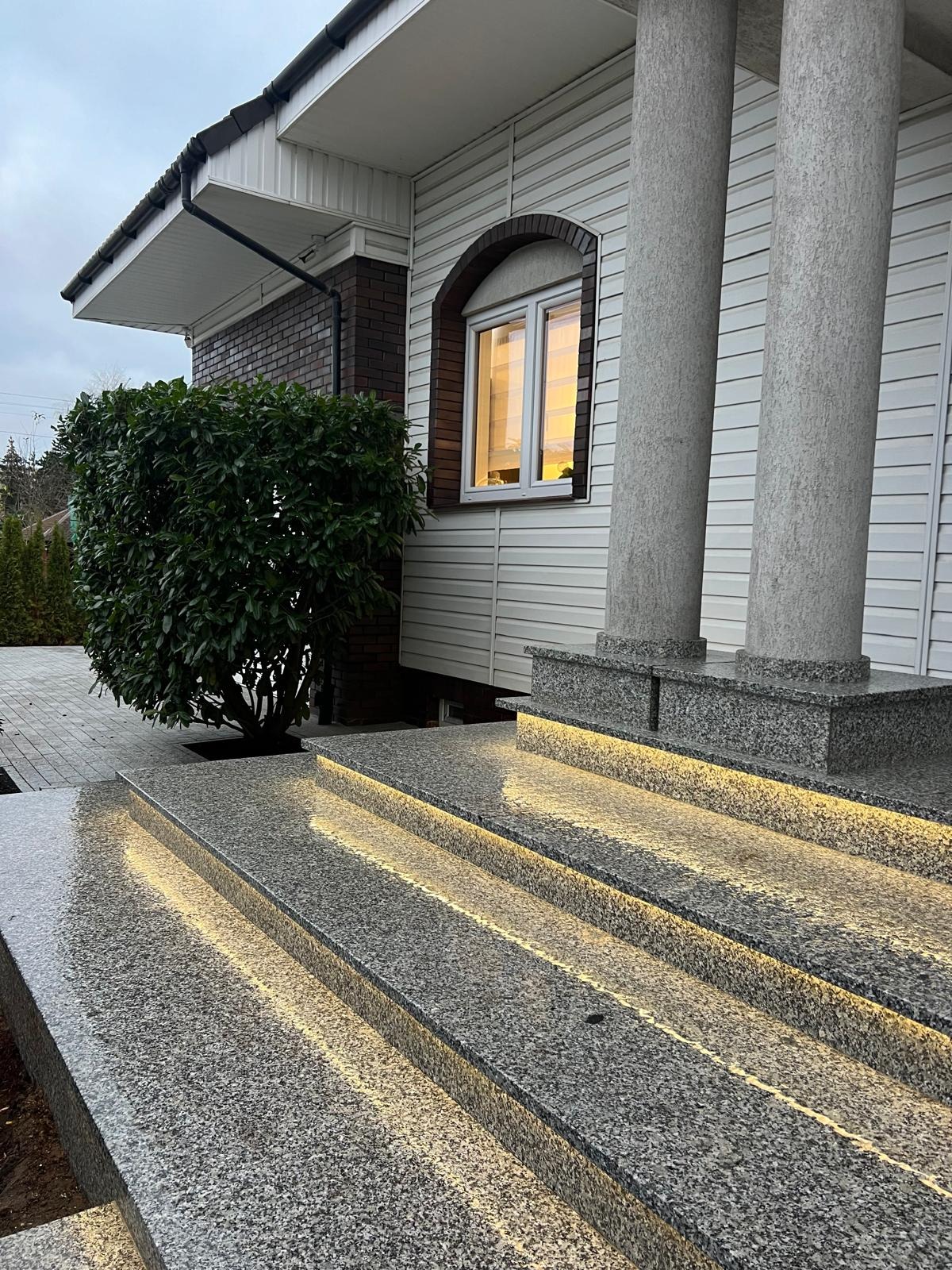 Granitowe schody zewnętrzne z podświetleniem LED prowadzące do domu z kolumnami. Widoczne detale wykończenia i oświetlenie podkreślające strukturę kamienia.