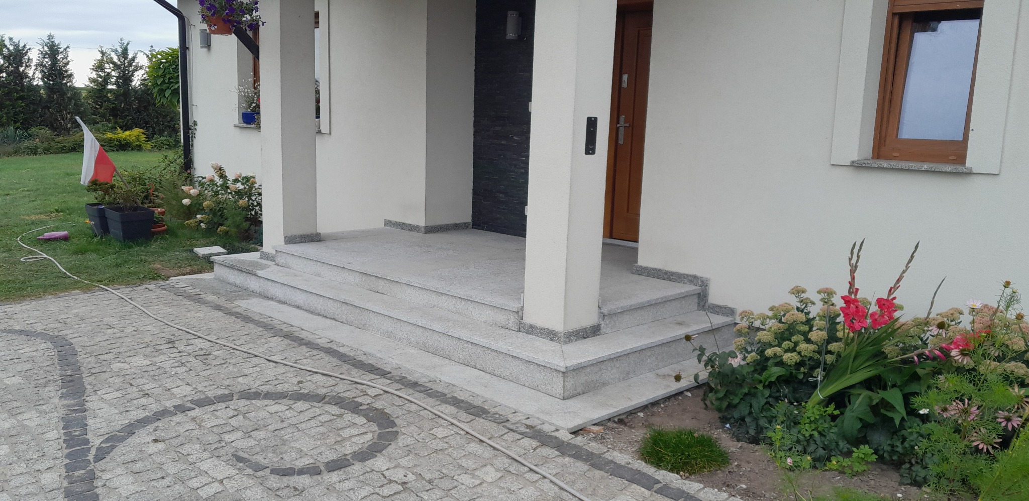 Granitowe schody wejściowe z szarej kostki brukowej prowadzące do domu z białymi ścianami i drewnianymi oknami, otoczone zielenią i kwiatami.