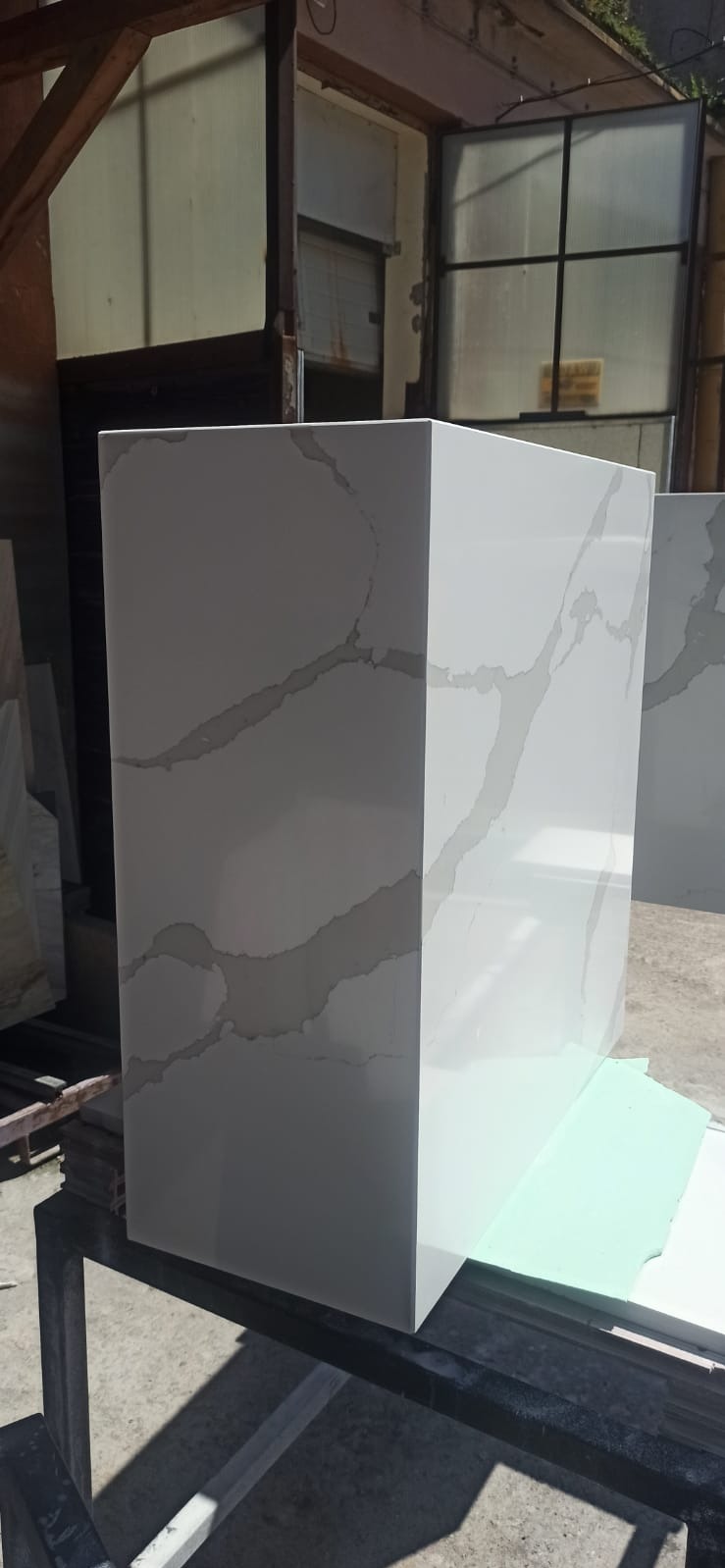 Biały, kwadratowy blok z szarymi żyłkami, prawdopodobnie z kamienia lub konglomeratu, stoi na metalowym stole w warsztacie kamieniarskim. Widoczne okno w tle.