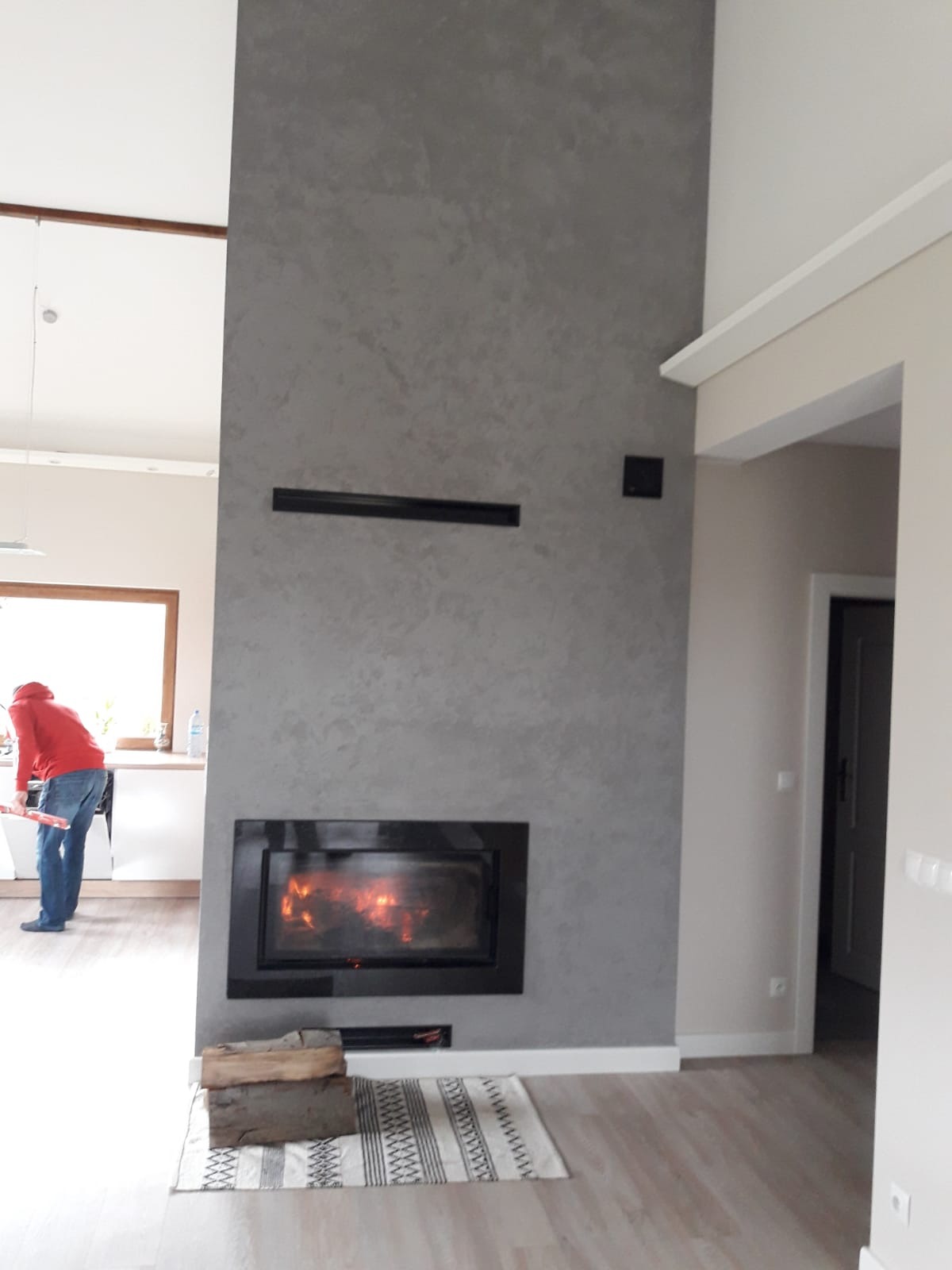 Nowoczesny kominek z szarą ścianą o fakturze betonu, płomienie w palenisku, drewno ułożone obok. W tle widoczna osoba w kuchni, co dodaje wnętrzu życia.