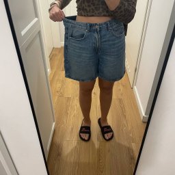 Tomasz Kapłun - Odbicie w lustrze kobiety trzymającej za duży rozmiar dżinsowych szortów, prezentującej utratę wagi. Widoczne klapki na stopach i fragment wnętrza.