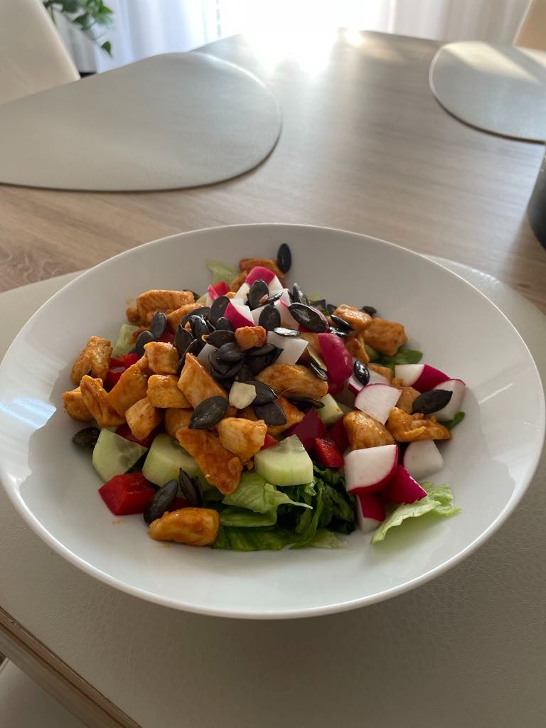 Kolorowa sałatka z kurczakiem, rzodkiewką, ogórkiem i pestkami dyni w białej misce na stole. Zdrowy posiłek pełen witamin, idealny dla osób dbających o linię.