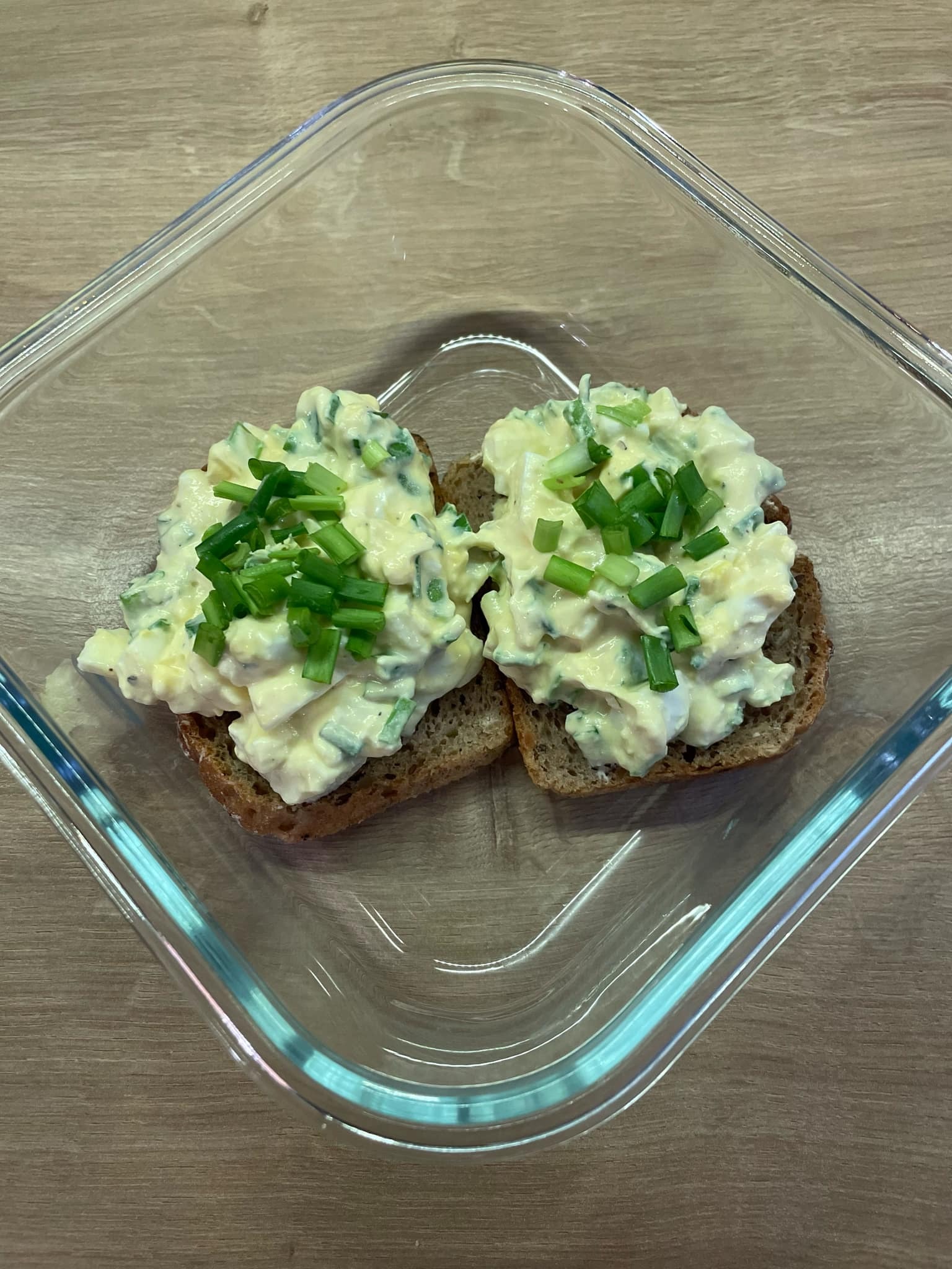 Dwie kromki ciemnego pieczywa z pastą jajeczną i szczypiorkiem w szklanym naczyniu na drewnianym stole, widok z góry, proste danie.