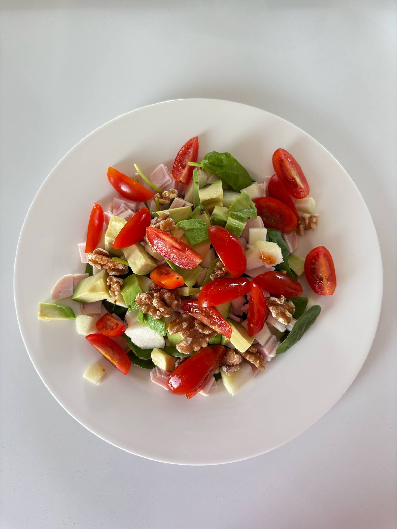 Kolorowa sałatka z pomidorkami, awokado, orzechami włoskimi i szynką na białym talerzu, w jasnym oświetleniu, z góry.