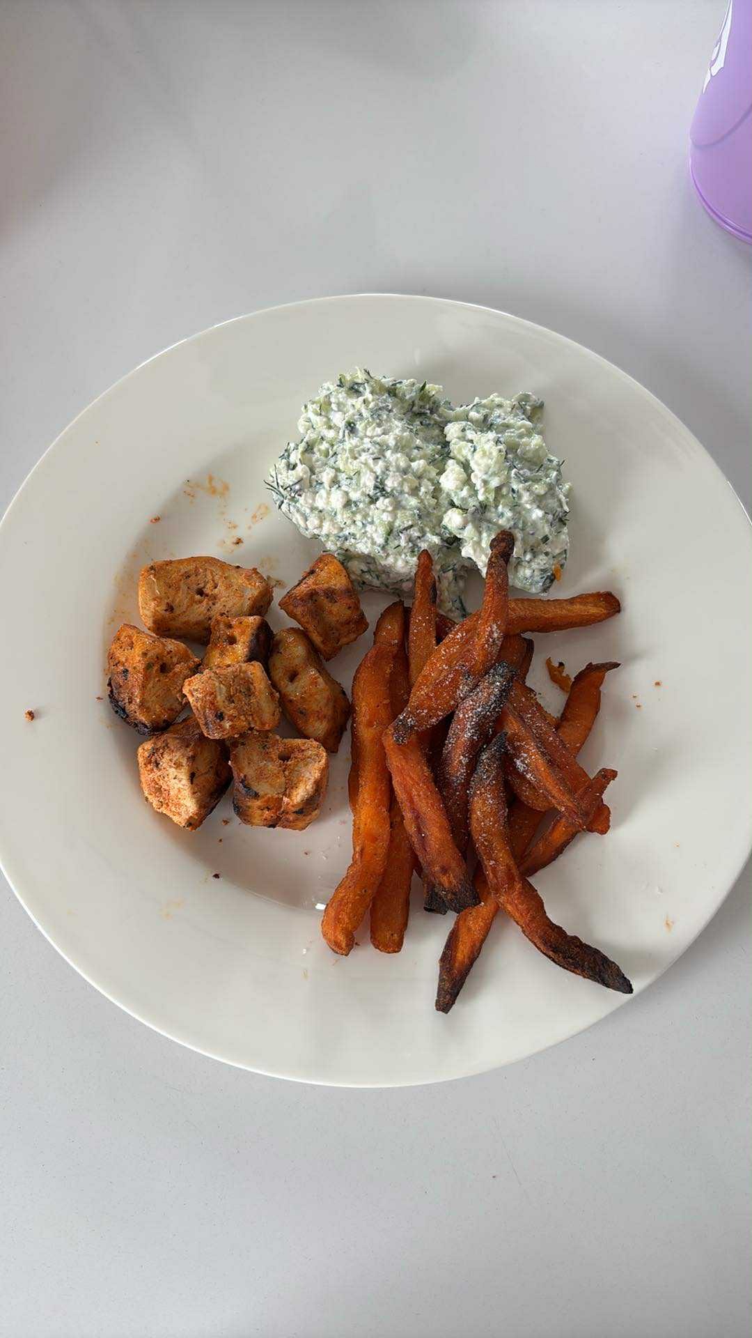 Talerz z grillowanym kurczakiem w kostkach, frytkami z batatów z przypalonymi końcówkami i sosem tzatziki na białym talerzu, na jasnym tle.