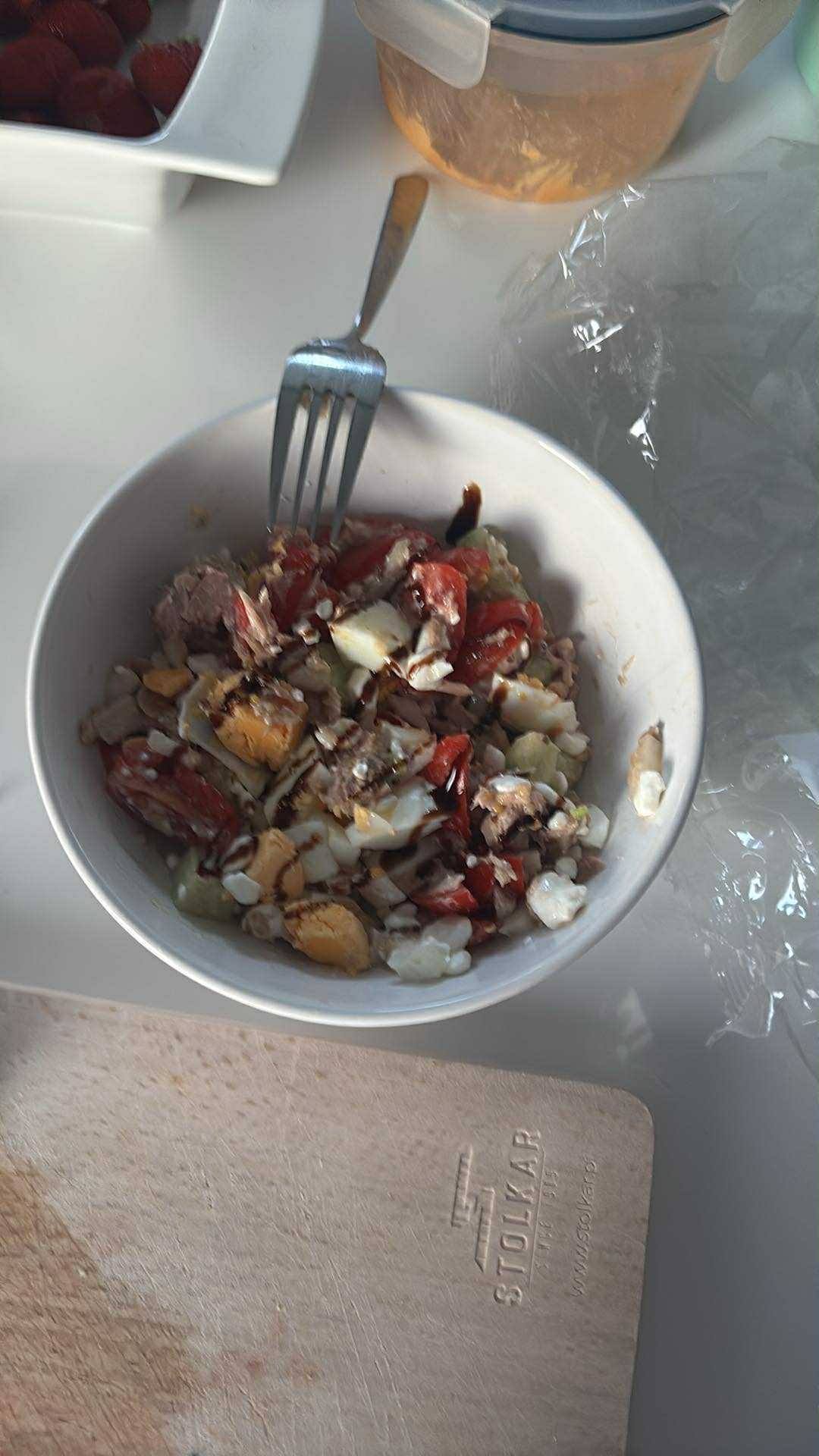 Sałatka z tuńczykiem, jajkiem i pomidorami w białej misce, z widelcem, na desce do krojenia Stolkar, obok pojemnik na jedzenie i truskawki.