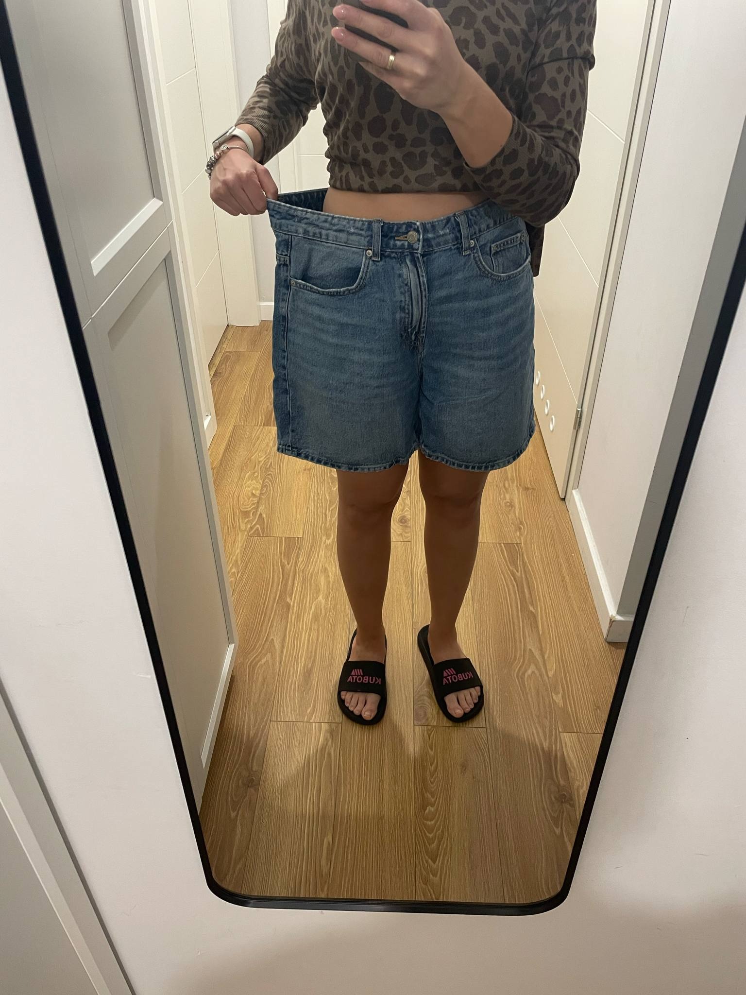 Odbicie w lustrze kobiety trzymającej za duży rozmiar dżinsowych szortów, prezentującej utratę wagi. Widoczne klapki na stopach i fragment wnętrza.