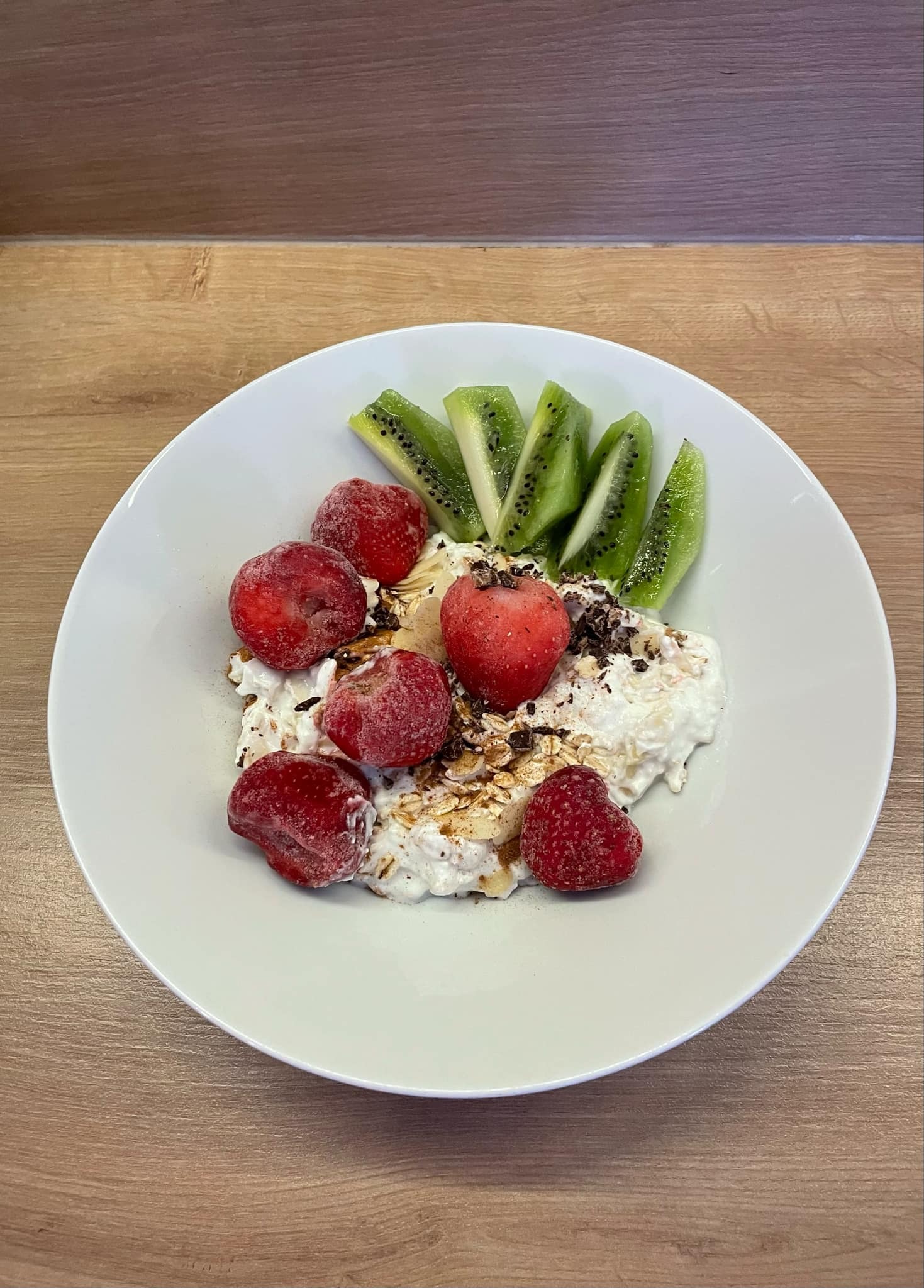Talerz z twarożkiem, owocami kiwi i mrożonymi truskawkami, posypany kawałkami czekolady na drewnianym stole. Zdrowy posiłek.