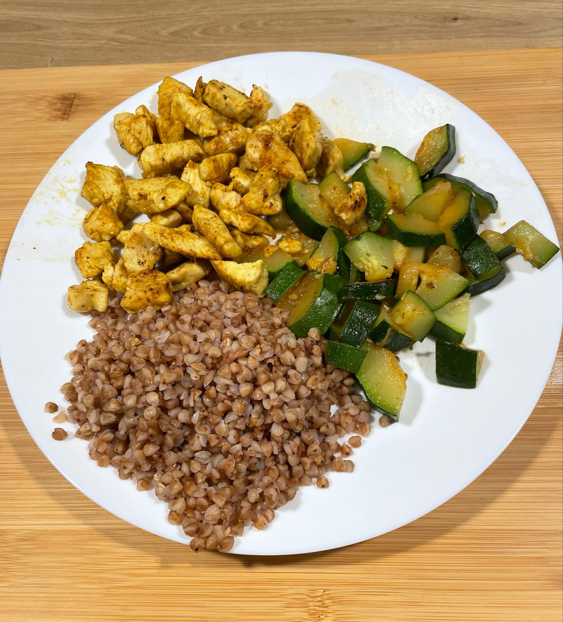 Talerz z porcją kaszy gryczanej, kurczaka w przyprawach i duszonej cukinii na drewnianym blacie. Zdjęcie z góry, ostre światło.