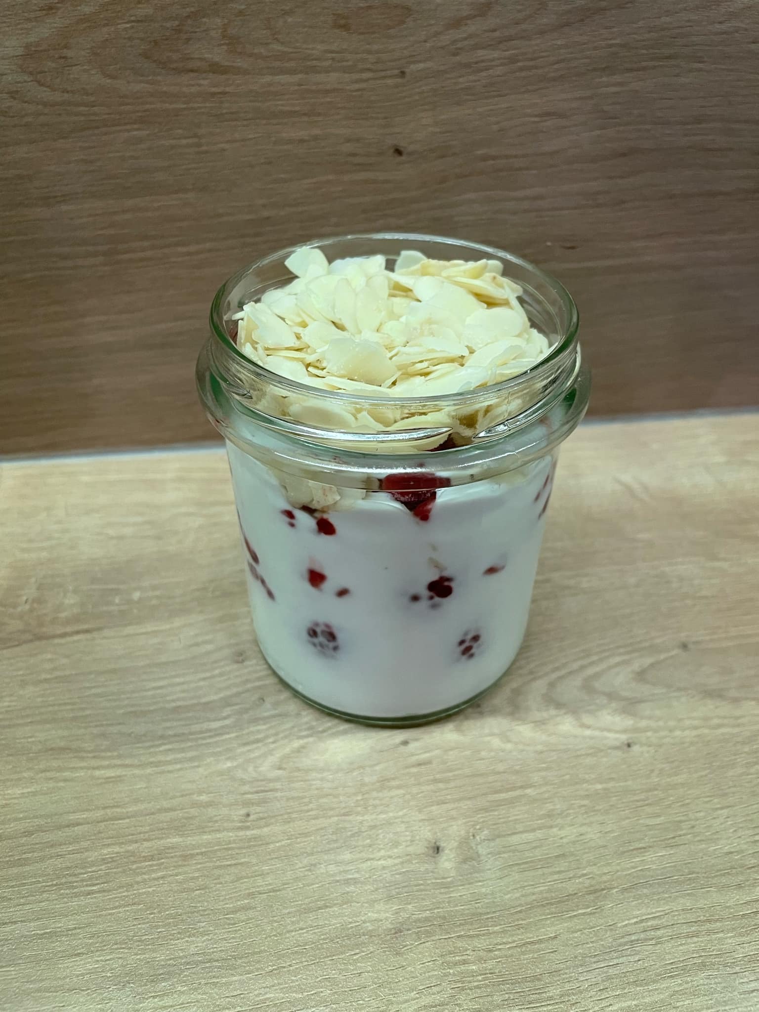 Deser w słoiku: jogurt z owocami i płatkami migdałów. Zdjęcie z bliska ukazuje warstwy i tekstury składników na drewnianym tle.