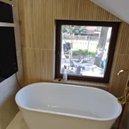 Biała wanna wolnostojąca w łazience na poddaszu z oknem i drewnianą boazerią. Widoczny wentylator sufitowy i złota armatura.