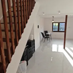 Jasne wnętrze po remoncie: drewniane schody, białe ściany, błyszcząca podłoga z płytek. Minimalistyczny wystrój, widok na jadalnię z oknem.