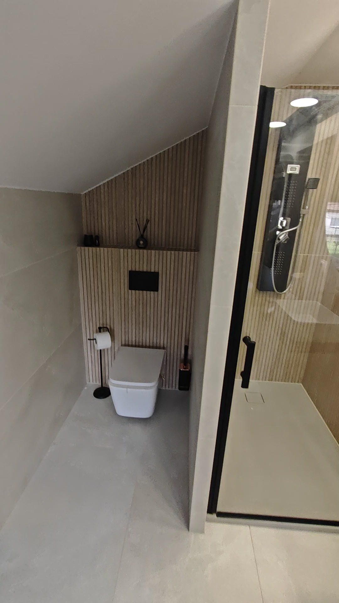 Nowoczesna łazienka na poddaszu ze ścianą z drewnianych lameli, kabiną prysznicową typu walk-in i podwieszaną toaletą. Minimalistyczny design z akcentami czerni.