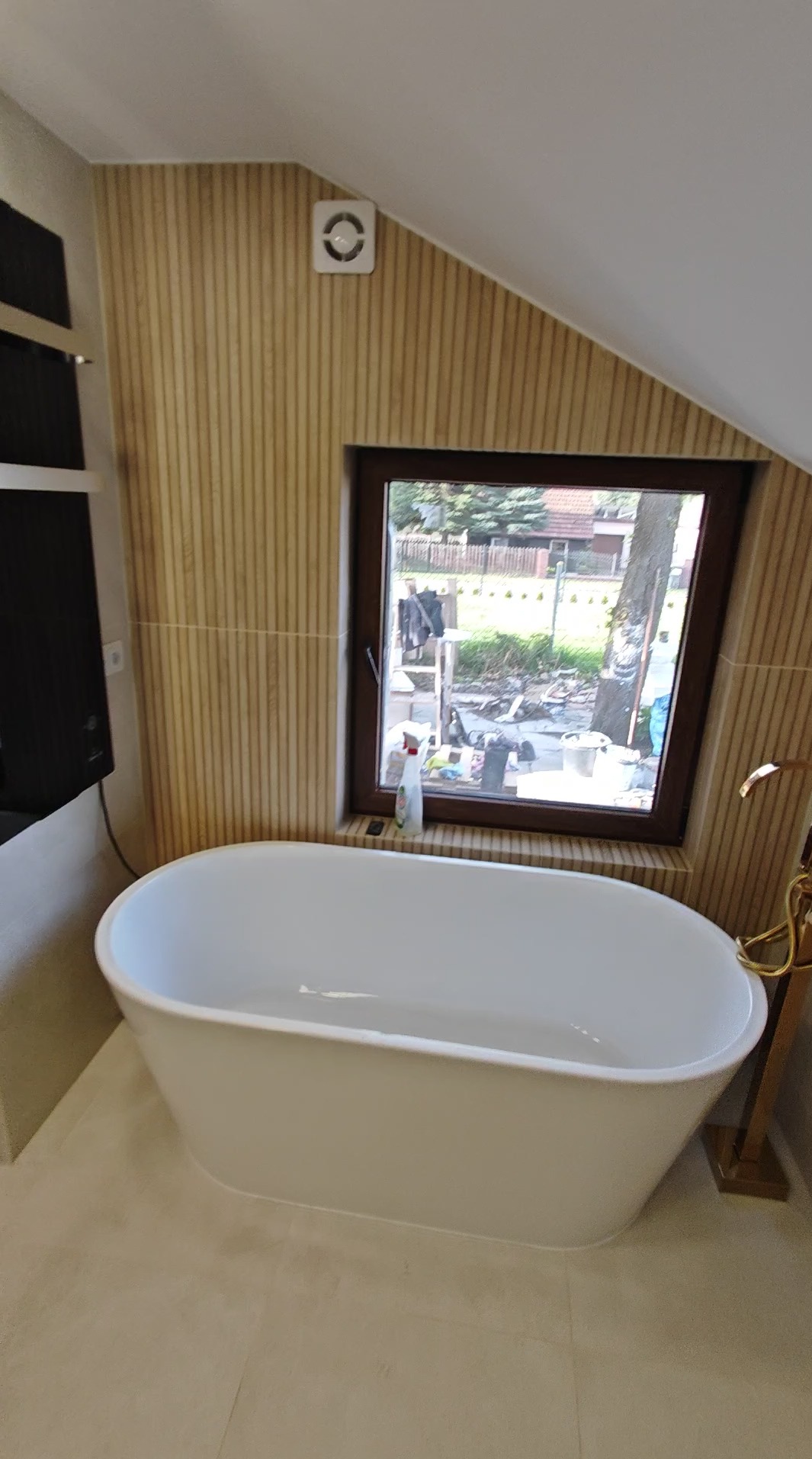 Biała wanna wolnostojąca w łazience na poddaszu z oknem i drewnianą boazerią. Widoczny wentylator sufitowy i złota armatura.