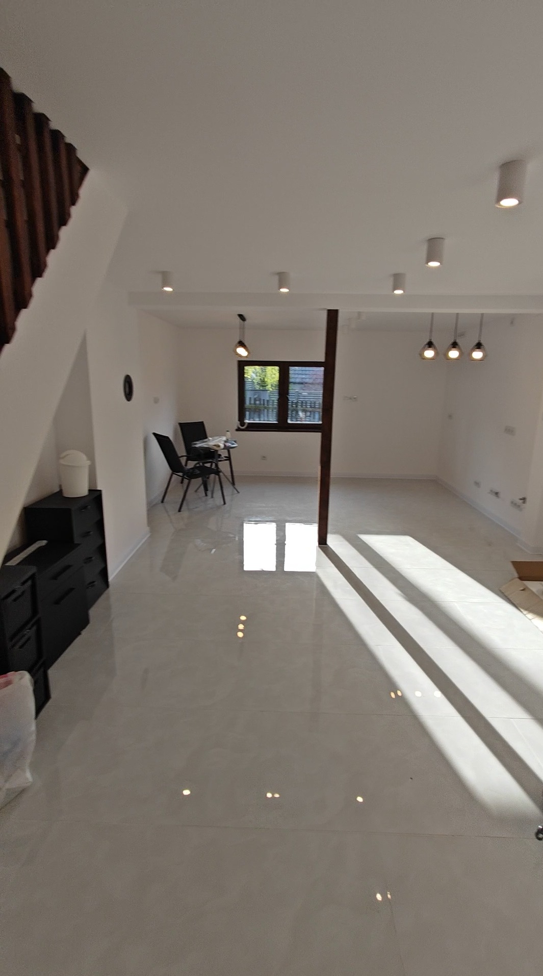 Jasne, przestronne wnętrze po remoncie z błyszczącą, kremową podłogą. Widoczne schody, okno z widokiem na zieleń i nowoczesne oświetlenie sufitowe.