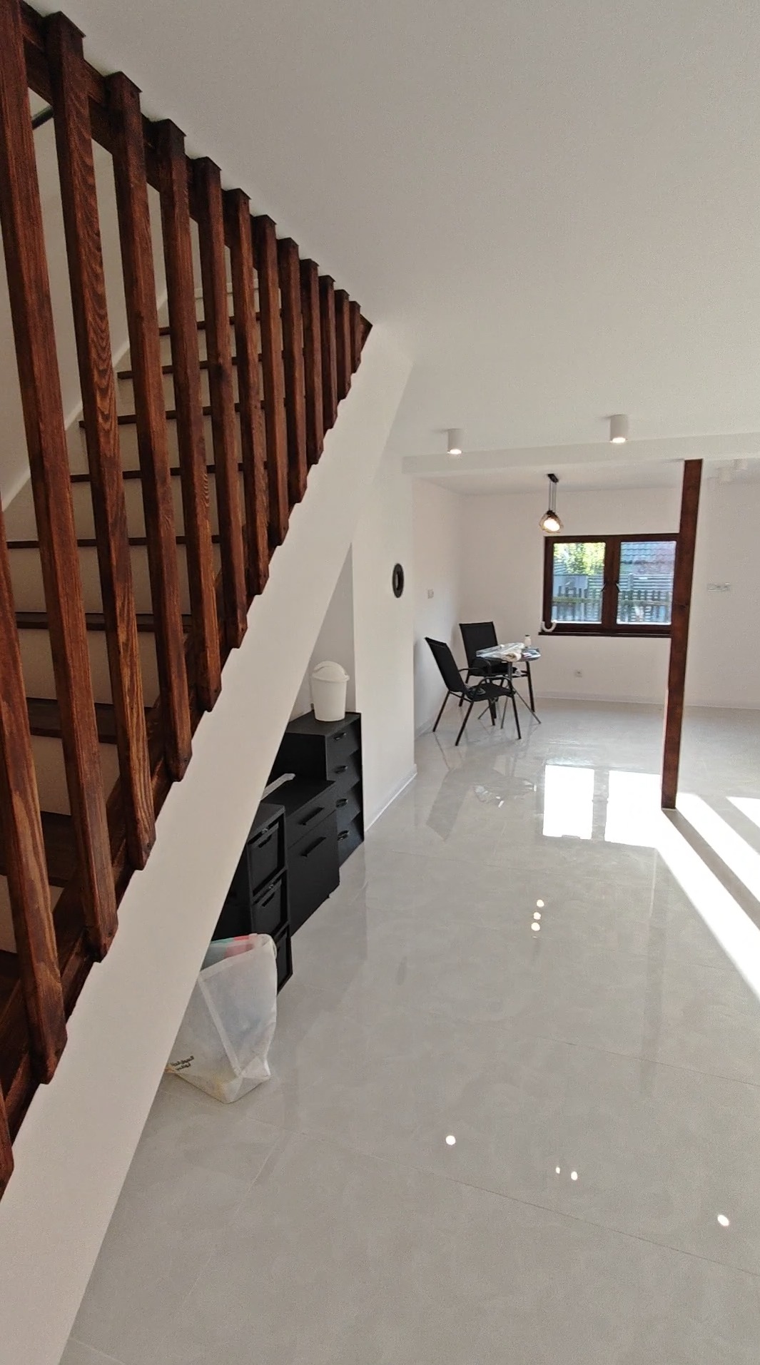 Jasne wnętrze po remoncie: drewniane schody, białe ściany, błyszcząca podłoga z płytek. Minimalistyczny wystrój, widok na jadalnię z oknem.