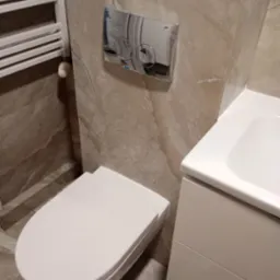 Wykończona łazienka z podwieszaną toaletą, płytkami imitującymi marmur i chromowanym przyciskiem spłukującym. Widoczne narzędzia i akcesoria glazurnicze.