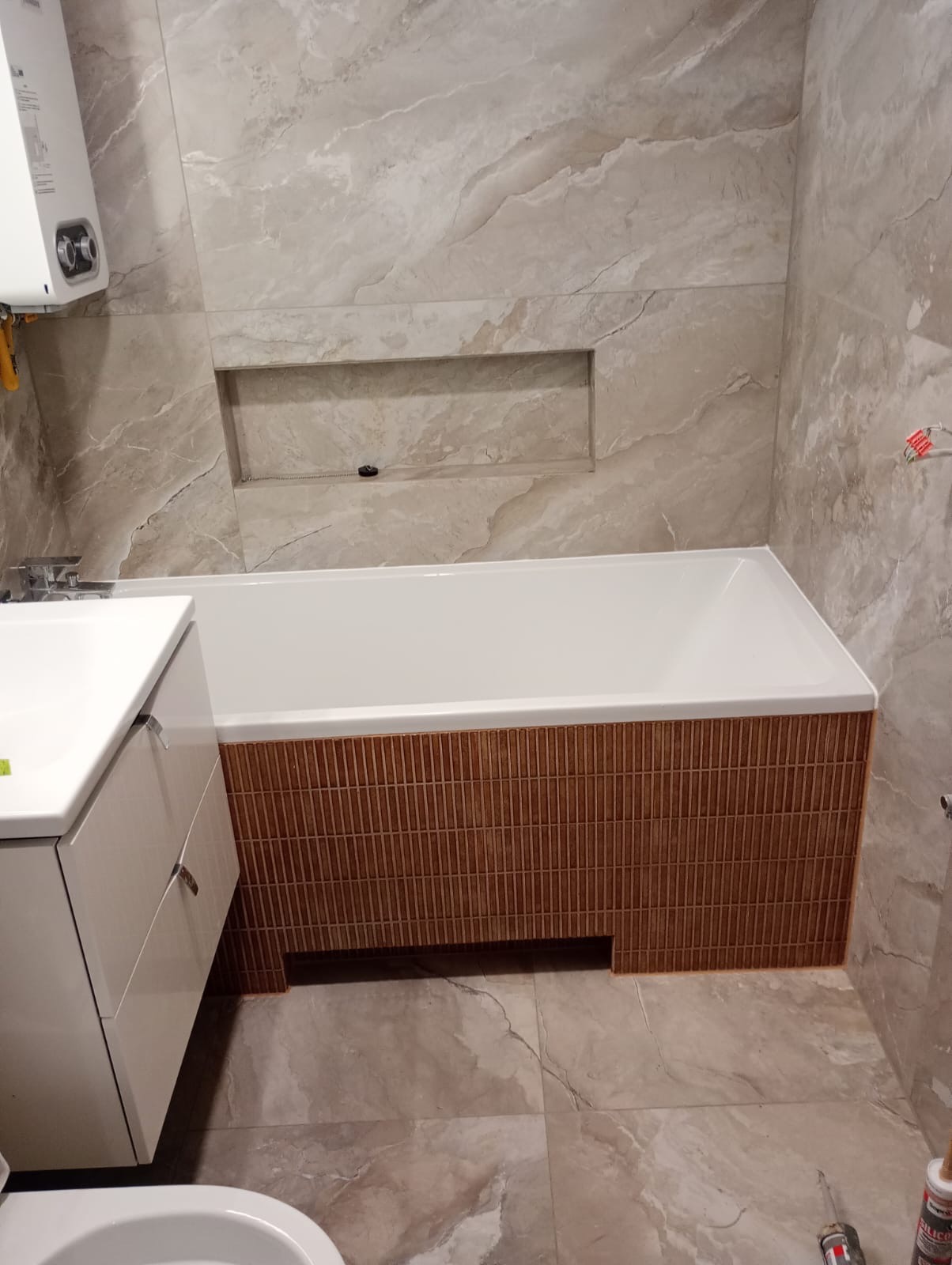 Wykończona łazienka: beżowe płytki imitujące marmur na ścianach i podłodze, zabudowana wanna z dekoracyjnymi, brązowymi płytkami, biała umywalka z szafką.