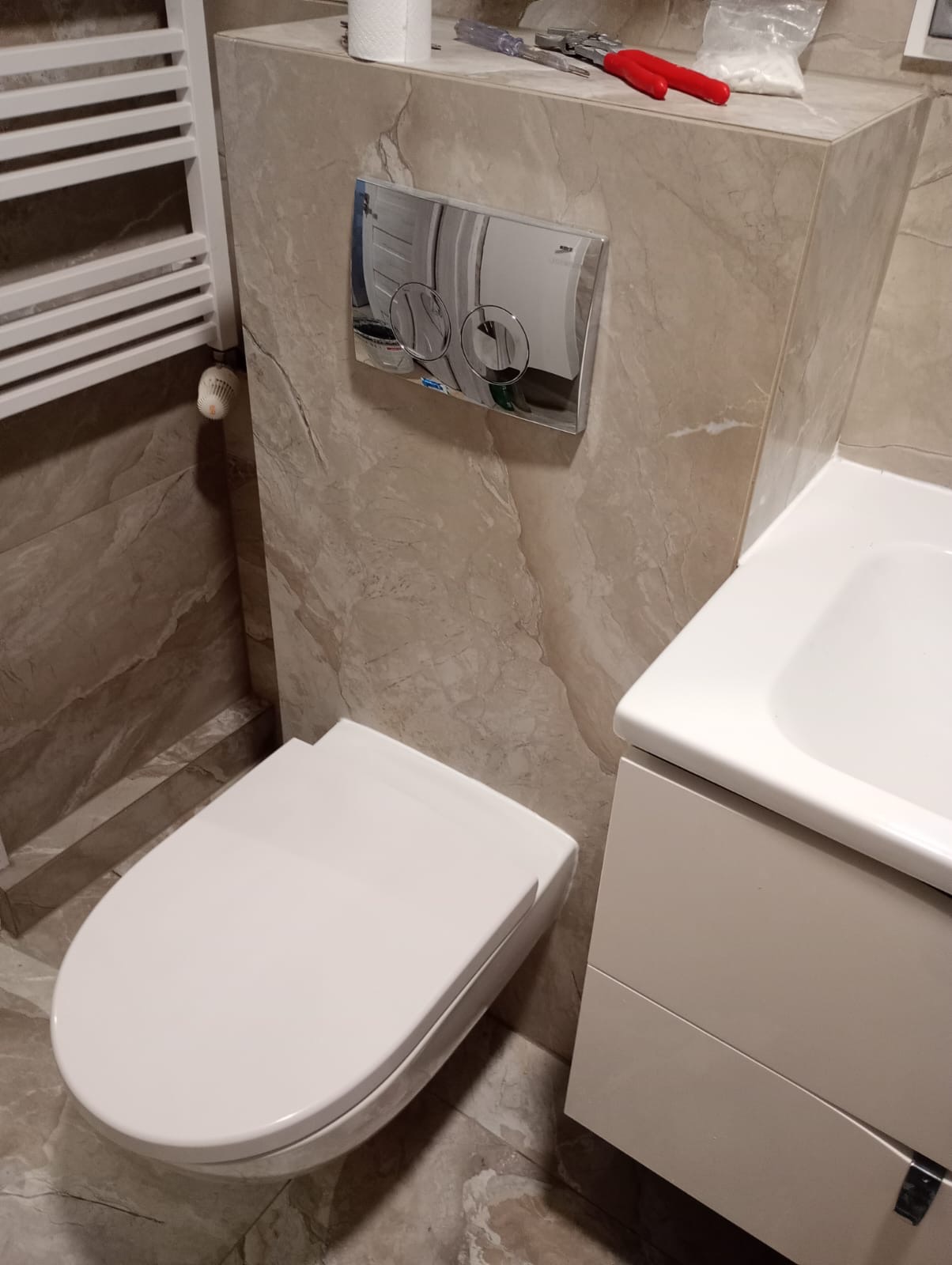 Wykończona łazienka z podwieszaną toaletą, płytkami imitującymi marmur i chromowanym przyciskiem spłukującym. Widoczne narzędzia i akcesoria glazurnicze.