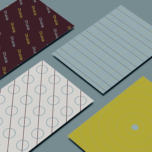 Cztery próbki druku o różnych wzorach i kolorach, ułożone pod kątem na szarym tle. Wzory obejmują paski, koła i logo 'Drukla'.