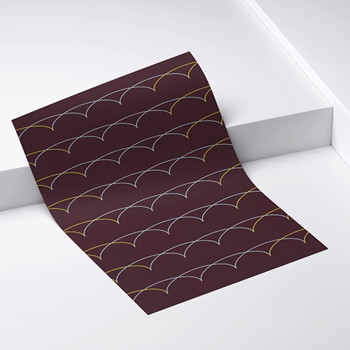 Brązowa kartka papieru z powtarzalnym wzorem łuków w kolorze złotym i białym, leżąca na białym tle. Układ ukośny, widoczne załamania papieru.