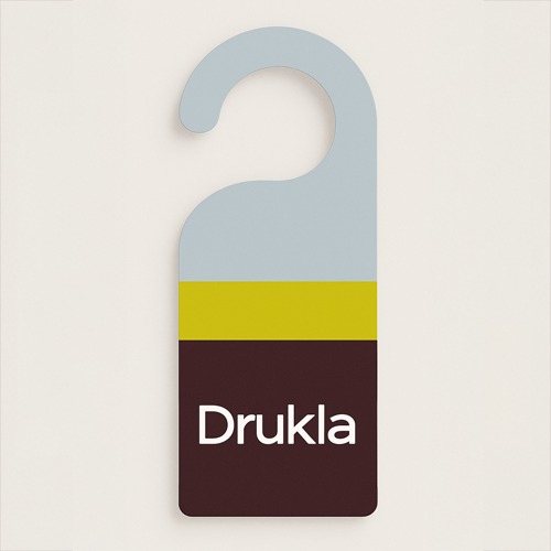 Kolorowy zawieszka na klamkę z napisem 'Drukla', w odcieniach szarości, brązu i żółci, na kremowym tle. Minimalistyczny design.