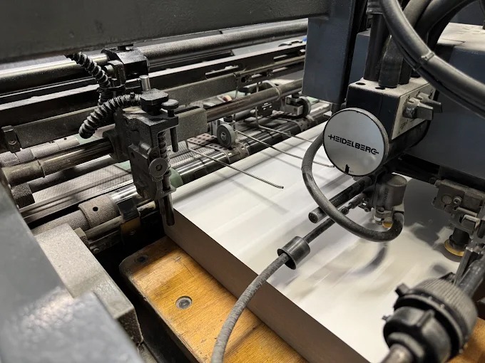 Proces druku offsetowego: arkusz papieru przechodzi przez maszynę Heidelberg, widoczne elementy mechaniczne i precyzyjne ustawienie.