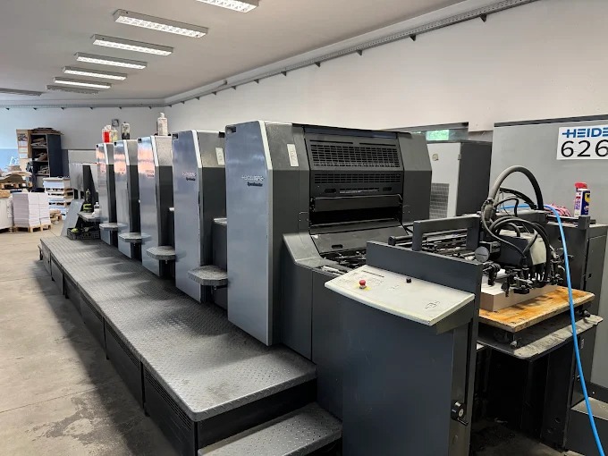 Duża, offsetowa maszyna drukarska Heidelberg Speedmaster w hali produkcyjnej. Widoczne podajniki papieru i panel sterowania. Oświetlenie jarzeniowe.